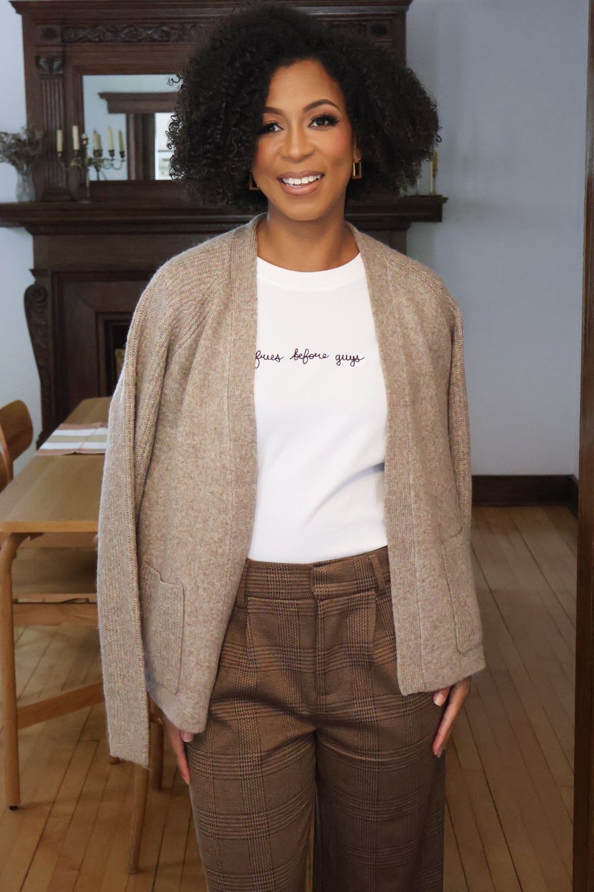 A woman with dark curly hair stands indoors wearing a beige textured cardigan over a white graphic T-shirt and brown plaid trousers. The background shows a wooden fireplace mantel and light wood floor.